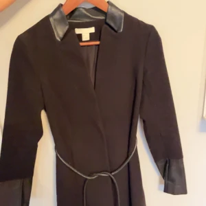 Svart kappa från H&M - Svart klänning från H&M med snygga detaljer i skinnimitation på krage och ärmslut. Klänningen har lång ärm och ett smalt bälte i midjan för en markerad siluett. Perfekt för dig som gillar stilrena och moderna plagg.