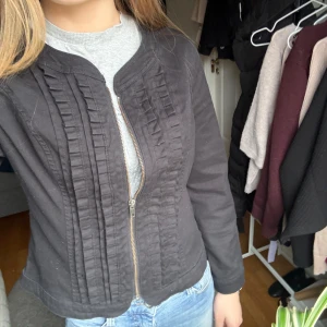 Svart jacka från Isolde - Säljer en svart ”jeans” jacka från Isolde med dekorativa volangdetaljer längs dragkedjan framtill. Jackan har rund halsringning och lång ärm, perfekt att styla till jeans eller kjol.