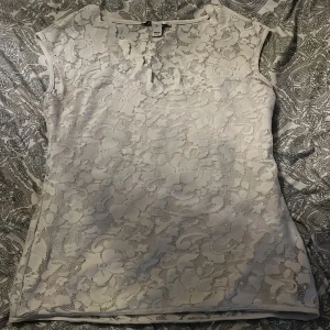 Vit spetsblus från NLY Trend - Säljer en vit blus från NLY Trend i storlek L. Blusen är ärmlös och har ett vackert blommigt spetsmönster över hela plagget med insytt under linne. Perfekt att styla med jeans eller kjol för en fräsch look.