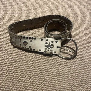 Silvrigt nitat bälte med dubbla spännen - Säljer ett vitt bälte i skinn med massor av nitar och dekorativa detaljer längs hela bältet. Bältet är i andvänt gott skick 