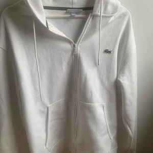 Vit hoodie från Lacoste med dragkedja - Säljer en klassisk vit hoodie från Lacoste med dragkedja framtill och två fickor. Använd fåtal gånger och bra skick!