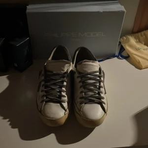 Vita Philippe Model sneakers med blå detalj - Säljer ett par vita Philippe Model sneakers med grå snörning och blå detalj på sidan. Skorna har en klassisk låg modell och platt sula, samt grå hälparti i kontrastmaterial. Perfekta för dig som gillar stilrena och trendiga sneakers.