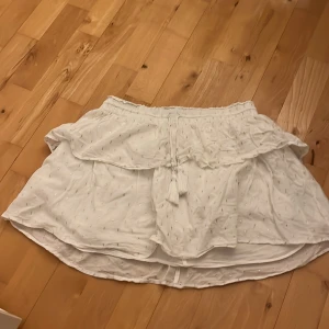Vit kjol med volang och knytband - Säljer en söt vit kjol med volang och knytband med tofsar i midjan. Kjolen har små glittriga detaljer och är tillverkad i ett lätt och luftigt material, perfekt för varma dagar.
