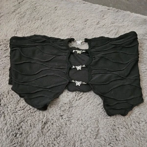 Svart tubtopp med dekorativa spännen - Säljer en svart tubtopp med vågiga sömmar och fjärilar på. Lappen finns inte kvar men tror den är från SHEIN vet heller inte vad det är för storlek men väldigt säker på att det är M men kan även passa en som har S