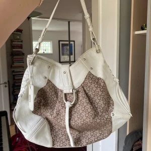 Skitsnygg väska från DKNY - Säljer en snygg axelväska från DKNY i vitt skinn och beige tyg med logomönster. Väskan har silverfärgade metalldetaljer. Perfekt för dig som vill ha en stilren och rymlig väska med klassisk design.☀️☀️❤️ Använd men i fint skick!