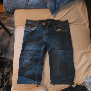 Blå jeansshorts från KappAhl - Säljer ett par klassiska blå jeansshorts från KappAhl med uppvikta benslut. Shortsen har fem fickor, knapp och dragkedja framtill samt synliga sömmar i kontrastfärg. Perfekta för varma dagar.