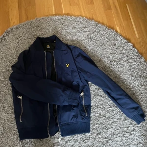 Marinblå bomberjacka från Lyle & Scott - Säljer en marinblå bomberjacka från Lyle & Scott med gul logga på bröstet. Jackan har dragkedja framtill, ribbade muddar och två fickor med dragkedja. Perfekt för dig som vill ha en stilren och klassisk look.