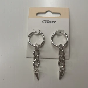 Silverfärgade örhängen med kedja och spets från Glitter - Säljer ett par coola silverfärgade örhängen från Glitter. De har en rund ring, en kedja och en spetsig detalj längst ner. Perfekta för att ge din look lite edge.