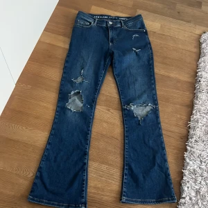Jättefina mörkblå lågmidjade bootcut jeans med slitningar - Säljer ett par blå bootcut jeans med tydliga slitningar och hål på benen. Jeansen har låg midja och klassisk femficksdesign. Perfekta för dig som gillar en avslappnad och trendig stil. Knappt använd 💗💗