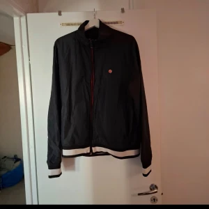 Björn Borg vårjacka - Säljer en svart vindjacka med vita och svarta ribbade muddar nertill och vid ärmslut. Jackan har dragkedja framtill och en liten röd logga på bröstet. Perfekt för svala dagar och enkel att matcha med det mesta.
