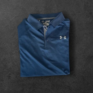 Blå golf piké från Under Armour - Snygg blå pikétröja från Under Armour med klassisk krage och knappar framtill. Tröjan har en vit logga på bröstet och är tillverkad i ett lätt och svalt material, perfekt för träning eller vardag. Passformen är loose för extra komfort. Golf piké.