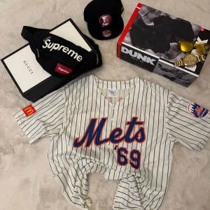 Randig Mets baseballskjorta - Säljer en vit baseballskjorta med blå tunna ränder och Mets-logga framtill. Skjortan har knappar hela vägen, korta ärmar och tryck med siffran 69 samt 'World Champs' på ryggen. Patchar på båda ärmarna. Perfekt för dig som gillar sportig stil.