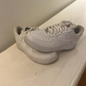 Vita Nike Air Force 1 sneakers - Klassiska helvita Nike Air Force 1 sneakers med snörning och rund tå. Skorna har en platt sula och är tillverkade i skinn med diskret Nike-logga på sidan och AIR-detalj på hälen. Perfekta för en clean och stilren look.