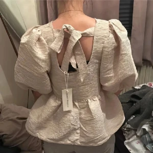 Beige puffärm blus med rosett i ryggen - Supersöt beige blus med puffiga ärmar och volang nertill. Baksidan har en snygg rosettdetalj som knyts i nacken. Blusen har en lätt strukturerad yta och är perfekt för dig som gillar romantisk stil. Helt ny med lappar❤️