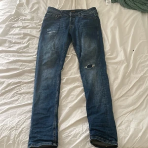 Blå slim fit jeans från Jack & Jones - Säljer ett par blå slim fit jeans från Jack & Jones, modell Glenn. Jeansen har slitna detaljer på låret och vid fickan, klassisk femficksdesign och knappgylf. Perfekta för dig som gillar en smalare passform och tidlös denimstil.