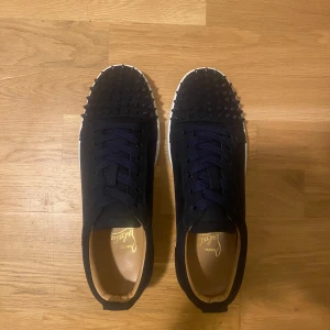 Mörkblåa sneakers med nitar från Christian Louboutin - Säljer ett par mörkblåa sneakers från Christian Louboutin med nitar på tån och klassisk röd sula. Skorna har snörning och rund tå, tillverkade i skinn och har en riktigt lyxig känsla.