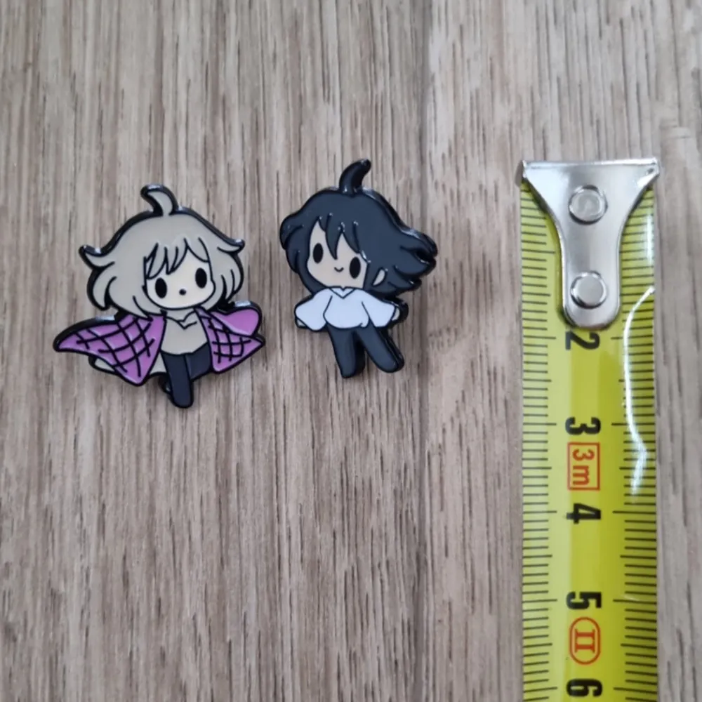 Två små pins föreställande animefigurer det levande slottet karaktärer pin, 2st. Den ena har ljust hår och en rosa-rutig mantel, medan den andra har mörkt hår och en vit tröja. Perfekta som accessoarer för att ge din outfit en unik touch!. Asusteet.