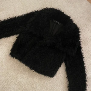 Svart pälsjacka från Zara - Säljer en snygg svart pälsjacka från Zara Basic. Den har långa ärmar och en klassisk krage. Perfekt för att ge din outfit en trendig touch. Finns ett hål i ena fickan annars är den som ny!!