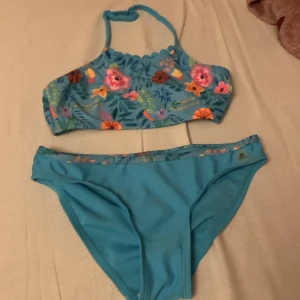 Blå blommig bikini  - Säljer en blå bikini med färgglatt blommigt mönster på både överdel och midjan på underdelen. Överdelen har hög hals och smala axelband. Perfekt för strand eller poolhäng! Den är storlek 140 men känns som 134.