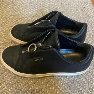 Svarta sneakers från Axel Arigato med dragkedja - Säljer ett par svarta sneakers från Axel Arigato med unik dragkedjedetalj och ring framtill istället för klassisk snörning. Storlek 36
