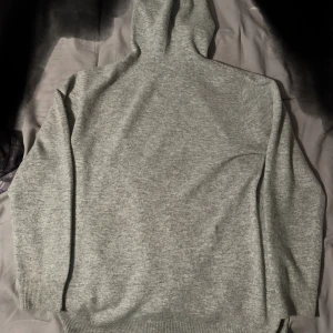 Ljusgrå stickad hoodie - Mysig Kashmir hoodie ljusgrå med huva och dragsko. Tröjan har långa ärmar och ribbade muddar vid ärmslut och nederkant. Perfekt för kyliga dagar och enkel att matcha med det mesta.