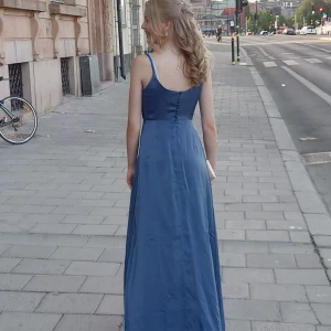 Blå långklänning med slits och knappar i ryggen - Säljer en elegant ljusblå långklänning med smala axelband, slits framtill och dekorativa knappar i ryggen. Klänningen har en mjuk, glansig yta och ett fint fall. Perfekt för bal eller festliga tillfällen. Inköpt på Bubbleroom. Använd endast en gång så klänningen är i nyskick. 