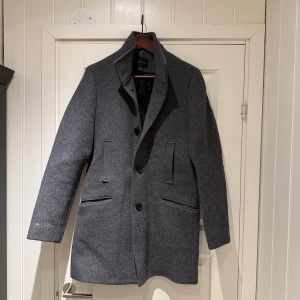 Grå ullkappa från Selected Homme - Säljer en stilren grå kappa från Selected Homme i ullblandning. Klassisk modell med knappar framtill, två fickor med lock och en snygg krage. Perfekt för dig som vill ha en tidlös och clean look.