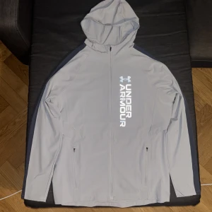 Ljusgrå hoodiejacka från Under Armour - Snygg ljusgrå hoodiejacka från Under Armour med dragkedja och huva. Jackan har svarta detaljer längs ärmarna och två fickor med dragkedja framtill. Stor logga och text på bröstet. Perfekt för träning eller chill.