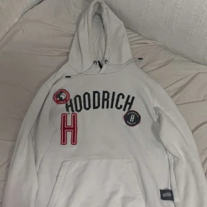 Vit hoodie från Hoodrich - Säljer en vit hoodie från Hoodrich med broderade märken och text på bröstet. Stor röd bokstav H och svarta bokstäver, samt två runda patches. Klassisk känguruficka och huva med dragsko.