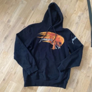 Svart Metallica hoodie med tryck - Cool svart hoodie med Metallica-tryck på bröstet och ryggen. Framsidan har en orange dödskalle med eldflammor och Metallica-loggan, medan ryggen har en dödskalle i en sol. Huva och känguruficka framtill.