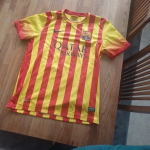 Barcelona fotbollströja med röd-gula ränder - Säljer en Barcelona fotbollströja från Nike med vertikala röd-gula ränder och Qatar Airways-tryck på bröstet. Tröjan har klubbmärke på vänster sida och korta ärmar med röd-gul färgövergång. Perfekt för dig som älskar Barca!