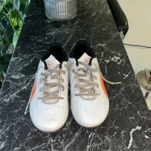 Vita fotbollsskor från Puma - Säljer ett par vita fotbollsskor från Puma med orange detaljer och snörning. Skorna har en rund tå och är designade för att ge bra grepp på planen med sina orange och svarta dobbar.
