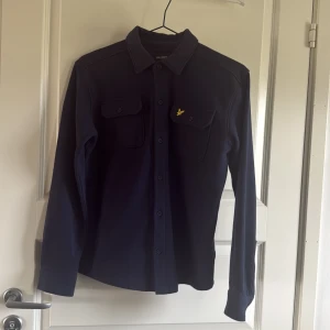 Mörkblå skjorta från Lyle & Scott - Snygg mörkblå skjorta från Lyle & Scott med klassisk krage och knappar framtill. Skjortan har två bröstfickor med lock och en gul broderad logotyp på ena fickan. Perfekt för en stilren look.