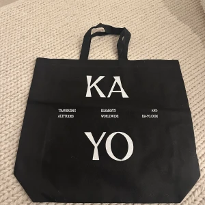 Svart shoppingväska från KA YO - Säljer en svart shoppingväska från KA YO med vita tryckta bokstäver och text på framsidan. Väskan har två handtag och är rymlig, perfekt för att bära med sig till vardags eller när du handlar.
