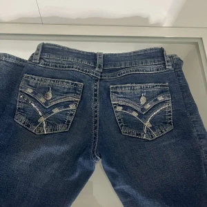 Blå jeansbyxor från Gina Tricot - Säljer ett par klassiska blå jeansbyxor från Gina Tricot med dekorativa bakfickor och tydliga kontrastsömmar. Jeansen har normal passform och raka ben, samt knapp och dragkedja framtill.