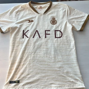 Vit fotbollströja Al Nassr Mané 10 - Snygg vit fotbollströja från Al Nassr med bruna detaljer och brunt tryck. Tröjan har korta ärmar, brunt ribbstickat avslut vid hals och ärmslut samt tryck med Mané och nummer 10 på ryggen. Perfekt för fotbollsfans!