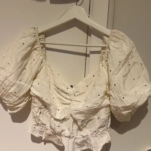 Vit blus med puffärmar från Gina Tricot - Supersöt vit blus med svarta prickar och stora puffärmar från Gina Tricot. Blusen har v-ringning, smockad rygg och volangdetaljer nertill. Perfekt för dig som gillar romantisk och trendig stil!