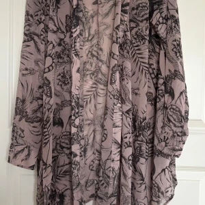 Ljusrosa mönstrad långärmad klänning - Säljer en ljusrosa kimono med svart botaniskt och djurinspirerat mönster. Klänningen har långa ärmar och en lös passform. Materialet känns lätt och luftigt, perfekt för vår och sommar. Snygg detalj med knappar i sidan.
