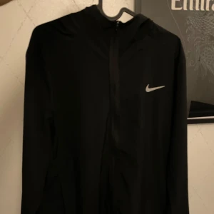Svart vindjacka från Nike Dri-FIT - Svart vindjacka från Nike med Dri-FIT-teknologi. Jackan har dragkedja framtill, huva och en diskret Nike-logga på bröstet. Perfekt för träning utomhus när det blåser. Jättesällan använd och i nyskick. Nypris 999kr
