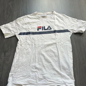 Vit t-shirt från Fila - Säljer en vit t-shirt från Fila med ett klassiskt tryck i rött och blått på framsidan. T-shirten har en normal passform och är kortärmad. Tröjan är knappt använd och märket har inga defekter 
