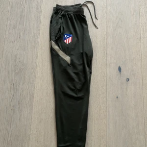 Mörkgröna träningsbyxor från Nike med Atlético Madrid-logga - Säljer ett par mörkgröna träningsbyxor från Nike med Atlético Madrid-logga på benet. Byxorna har dragkedja vid fickan, snörning i midjan och en beige detalj längs sidan. Storlek M men sitter mer som S