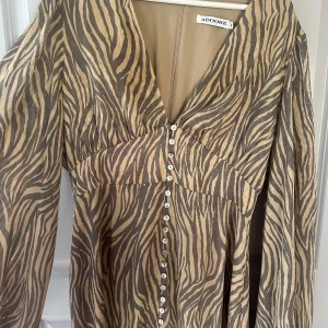 Zebramönstrad långklänning från Adoore - Säljer en långklänning från Adoore med zebramönster i beige och bruna toner. Klänningen har v-ringning, långa ärmar och knappar framtill. Helt perfekt klänning som jag tyvärr knappt använt! Är därför i väldigt fint skick. 