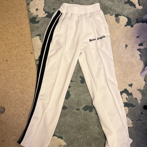 Vita byxor från Palm Angels med svarta ränder - Säljer ett par vita byxor från Palm Angels med svarta ränder längs sidorna och elastisk midja. Byxorna har rak passform och logga broderad framtill. Perfekta för en sportig och trendig look.