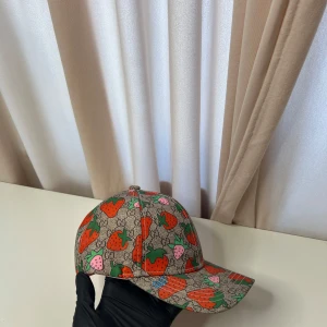 Gucci jordgubbs keps - Snygg keps från Gucci med beige bas och färgglatt jordgubbsmönster i rött, grönt och rosa. Kepsen har böjd skärm och justerbart rosa band bak. Perfekt för dig som vill sticka ut med en unik accessoar.
