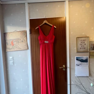 Röd/orange/corall balklänning med strassdetalj - Säljer en röd långklänning med djup v-ringning och smala axelband. Klänningen har en snygg strassdetalj i midjan som ger extra glamour. Perfekt för bal eller festliga tillfällen. Materialet känns lätt och faller fint. Saknas en diamant men går enkelt att fixa genom att beställa en ny