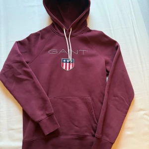 Vinröd hoodie från GANT - Säljer en vinröd hoodie från GANT med broderad logga på bröstet. Tröjan har huva med vita snören. Perfekt för många olika tillfällen. Använd fåtal gånger.