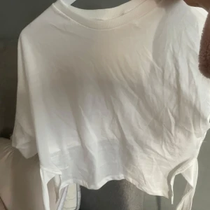 Vit croppad t-shirt med knyt - Säljer en vit croppad t-shirt med knyt i sidorna. T-shirten har rund halsringning och korta ärmar. Perfekt för en avslappnad och trendig stil.