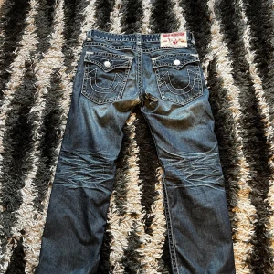 True Religion jeans - Säljer ett par feta True Religion Joey Super T jeans med tydliga kontrastsömmar och stora bakfickor med lock och knappar. Jeansen har professionellt lagats (se bild 4) och det syns knappt när man har dem på. Lägg gärna ett bud