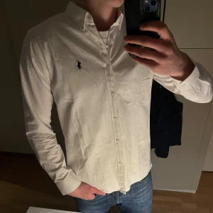 Vit skjorta från Ralph Lauren - Klassisk vit skjorta från Ralph Lauren med broderad logga på bröstet. Skjortan har lång ärm, knappar framtill och en stilren krage. Perfekt för dig som gillar en tidlös och clean look.