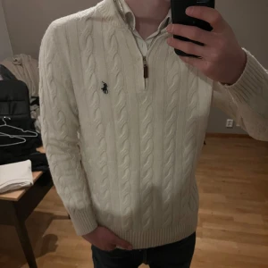 Vit kabelstickad tröja med dragkedja från Ralph Lauren - Säljer en klassisk vit kabelstickad tröja från Ralph Lauren med halv dragkedja och broderad logga på bröstet. Tröjan har lång ärm och ribbade muddar. Perfekt för lager-på-lager och stilren look.
